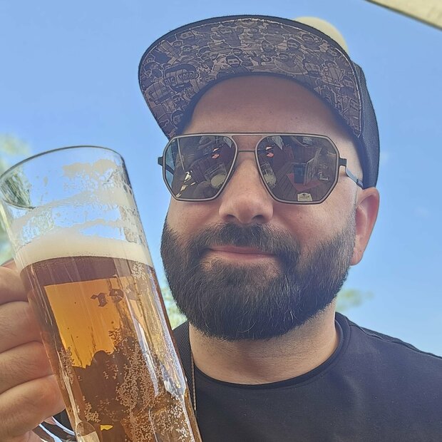Dr Emkus Junger Mann mit Bart, Sonnenbrille und Basecap schaut großes Bierglas an