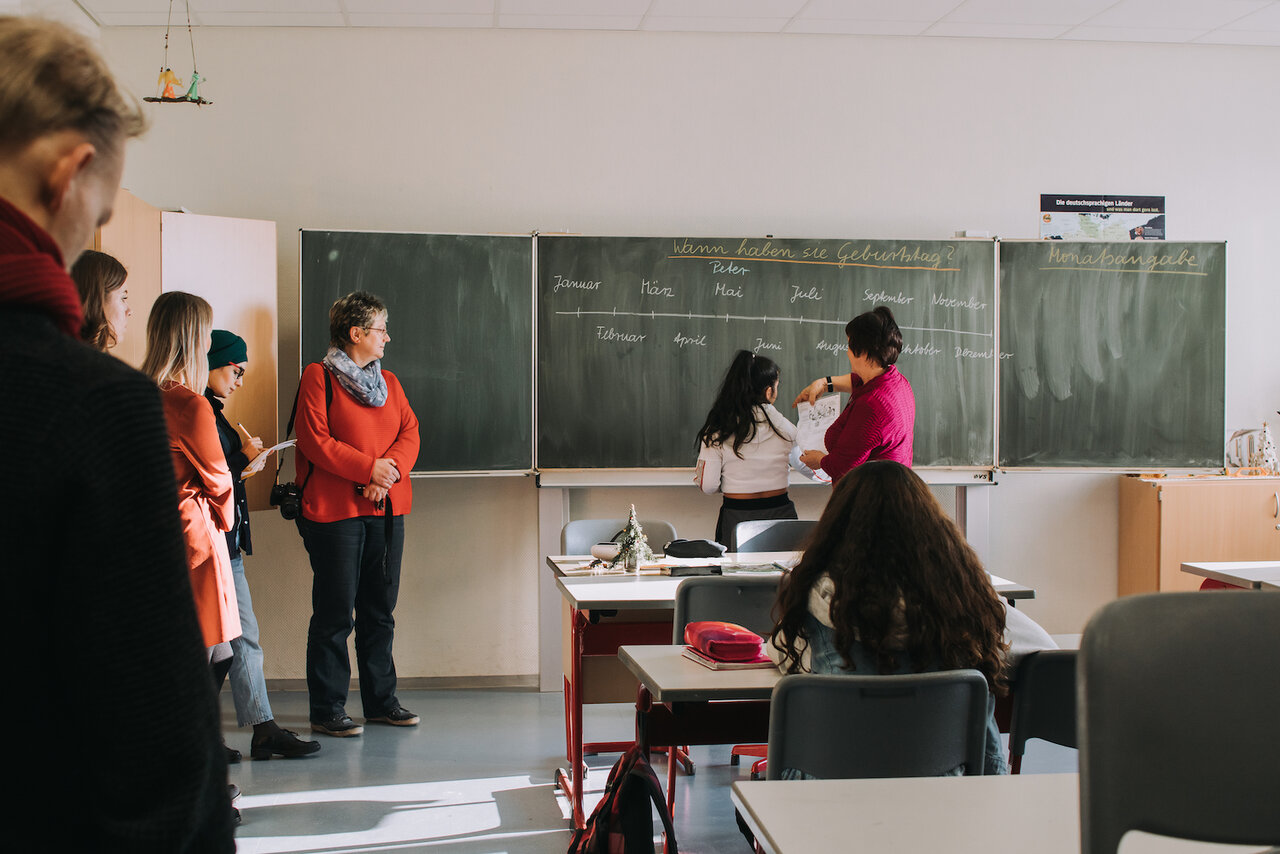 Klassenzimmer mit Blick auf Tafel, mehrere Personen stehen an der Seite