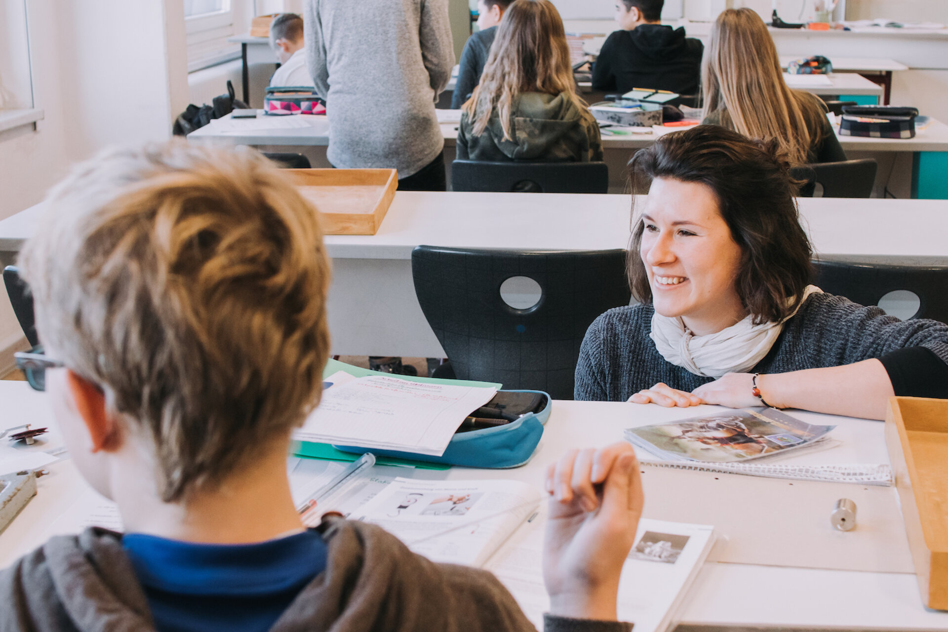 Blondes Kind von hinten, dunkelhaarige Lehrerin lehnt sich auf die Schulbank